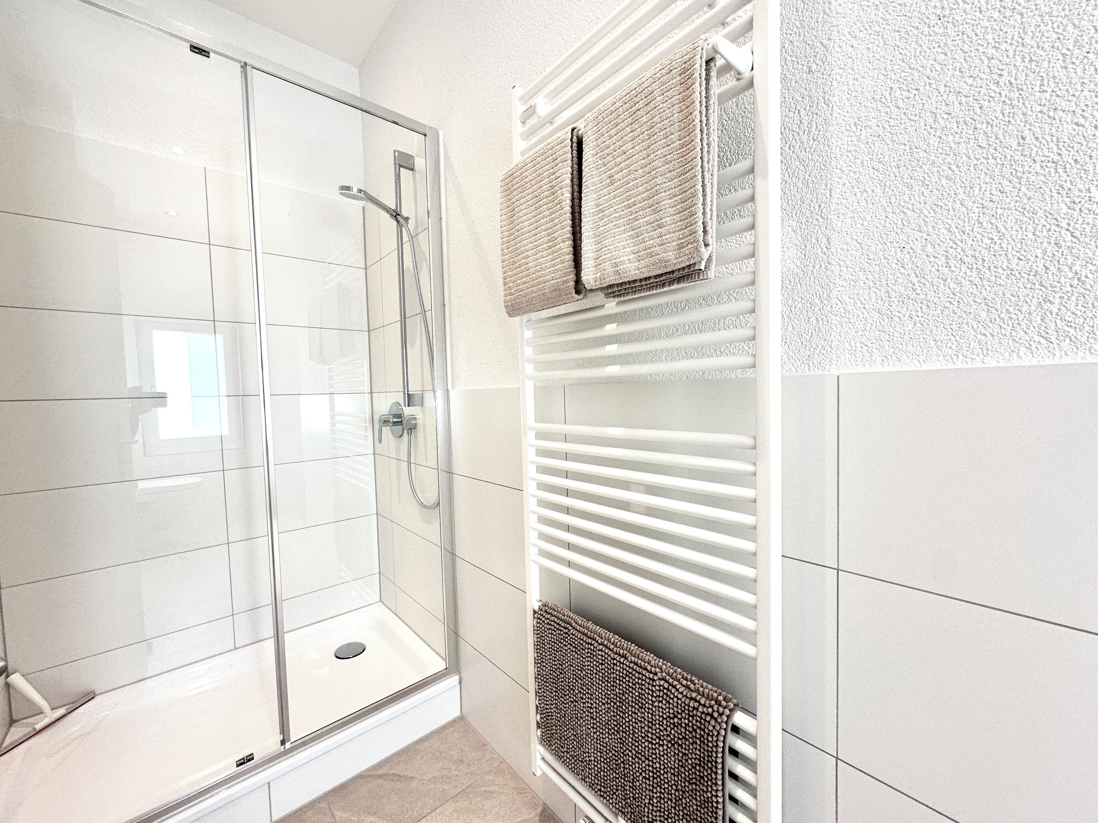 Une salle de bain moderne dotée d’une douche à parois de verre, de murs recouverts de carreaux blancs et d’un porte-serviettes intégré avec deux serviettes.