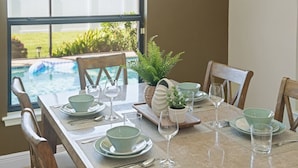 A dining table set for six with green plates and glasses, a potted plant, and a view of a swimming pool.