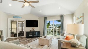 Un salón espacioso con ventilador de techo, televisor de pantalla plana, un ventanal con vistas al exterior y un cartel que dice "BEACH HOUSE".