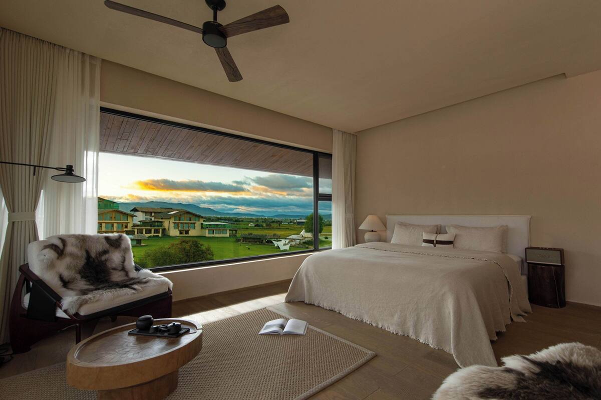 A bedroom with a large window, a bed, a chair, a bedside table, and a ceiling fan.