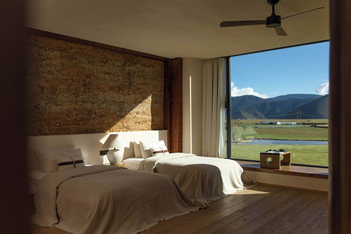 A bedroom with two beds, a large window with a view of mountains, and a ceiling fan.