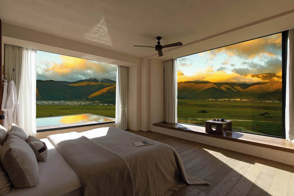A bedroom with a large bed, a ceiling fan, and a view of a mountainous landscape through large windows.