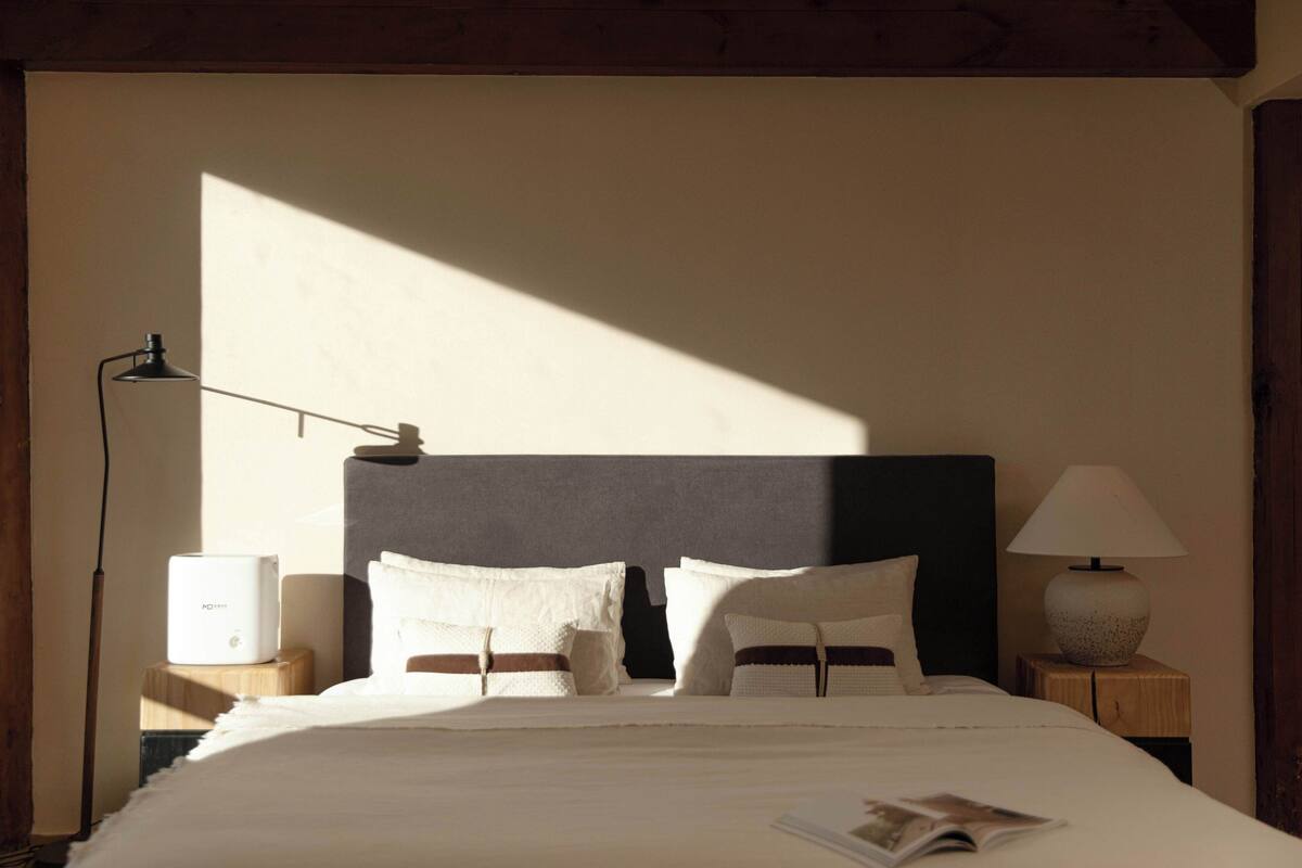 A neatly made bed with pillows and a magazine, a bedside lamp, and a small table with a lamp.