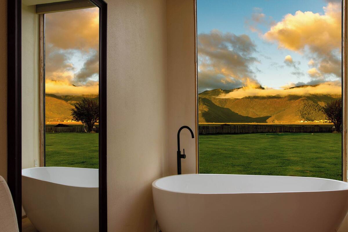 A bathroom with a bathtub, a window, and a view of a grassy field and hills.