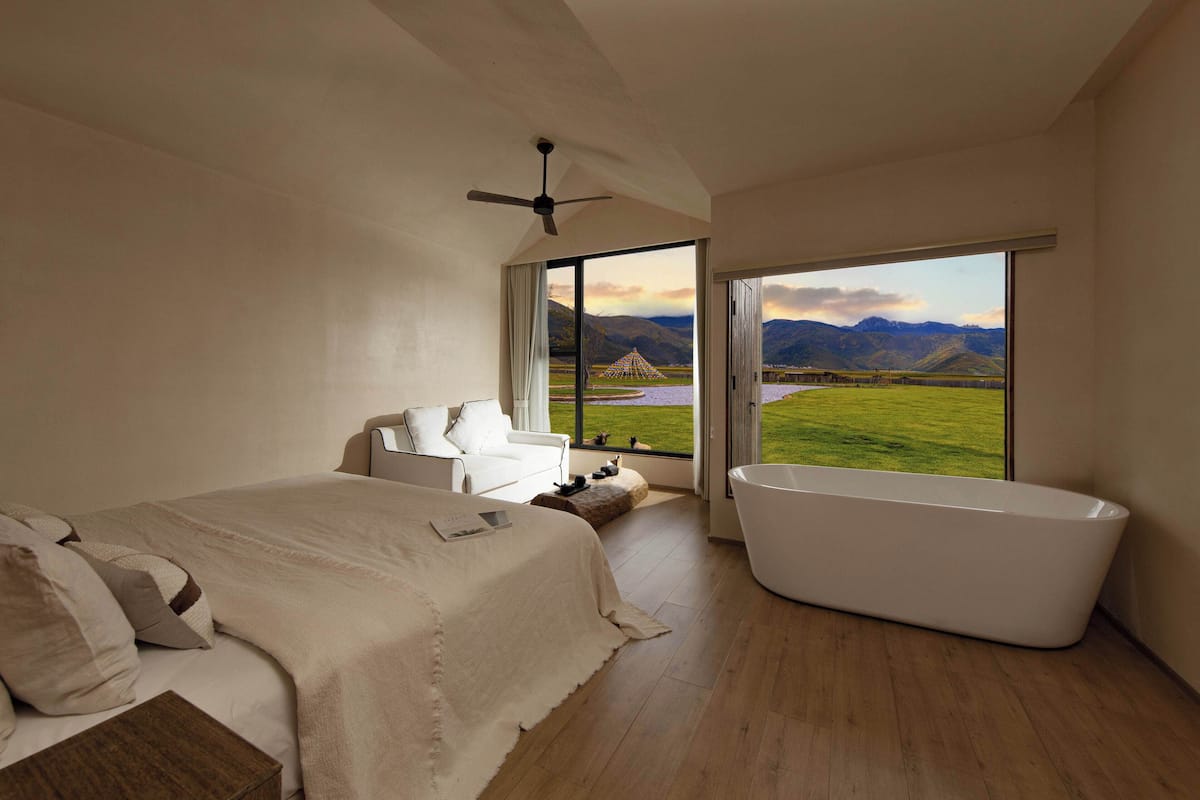 A bedroom with a large bed, a white bathtub, and a view of mountains and a lake.