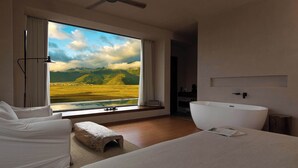 A modern hotel room with a large window offering a scenic view of mountains and a lake, a white bathtub, a ceiling fan, and a cozy seating area. - Liz's heart is in the sky (Deqin)