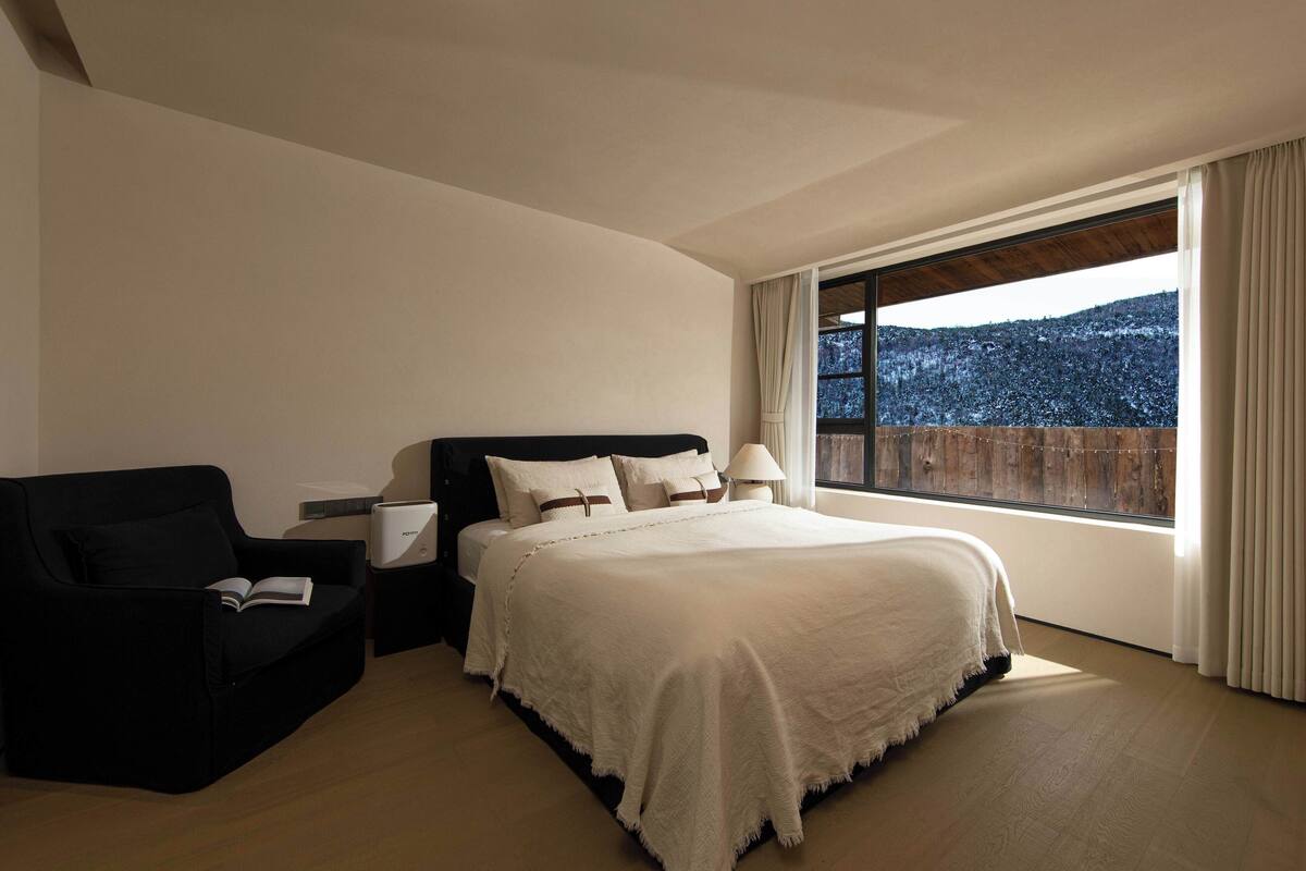 A bedroom with a bed, a black armchair, a bedside table, and a window with a view of a snowy landscape.