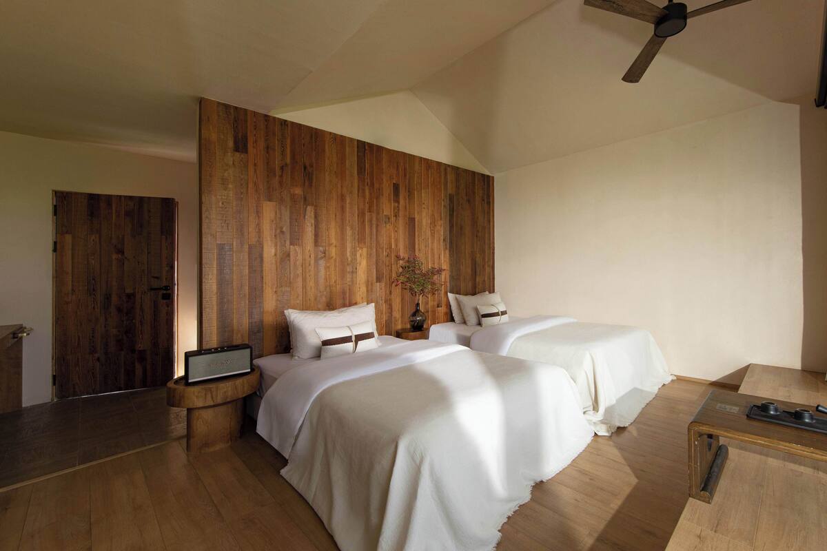 A modern bedroom with two beds, a wooden accent wall, a ceiling fan, and a bedside table with a laptop.