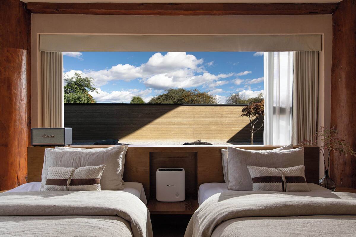 A bedroom with two beds, a bedside table, a bedside lamp, and a window with a view of the sky and trees.