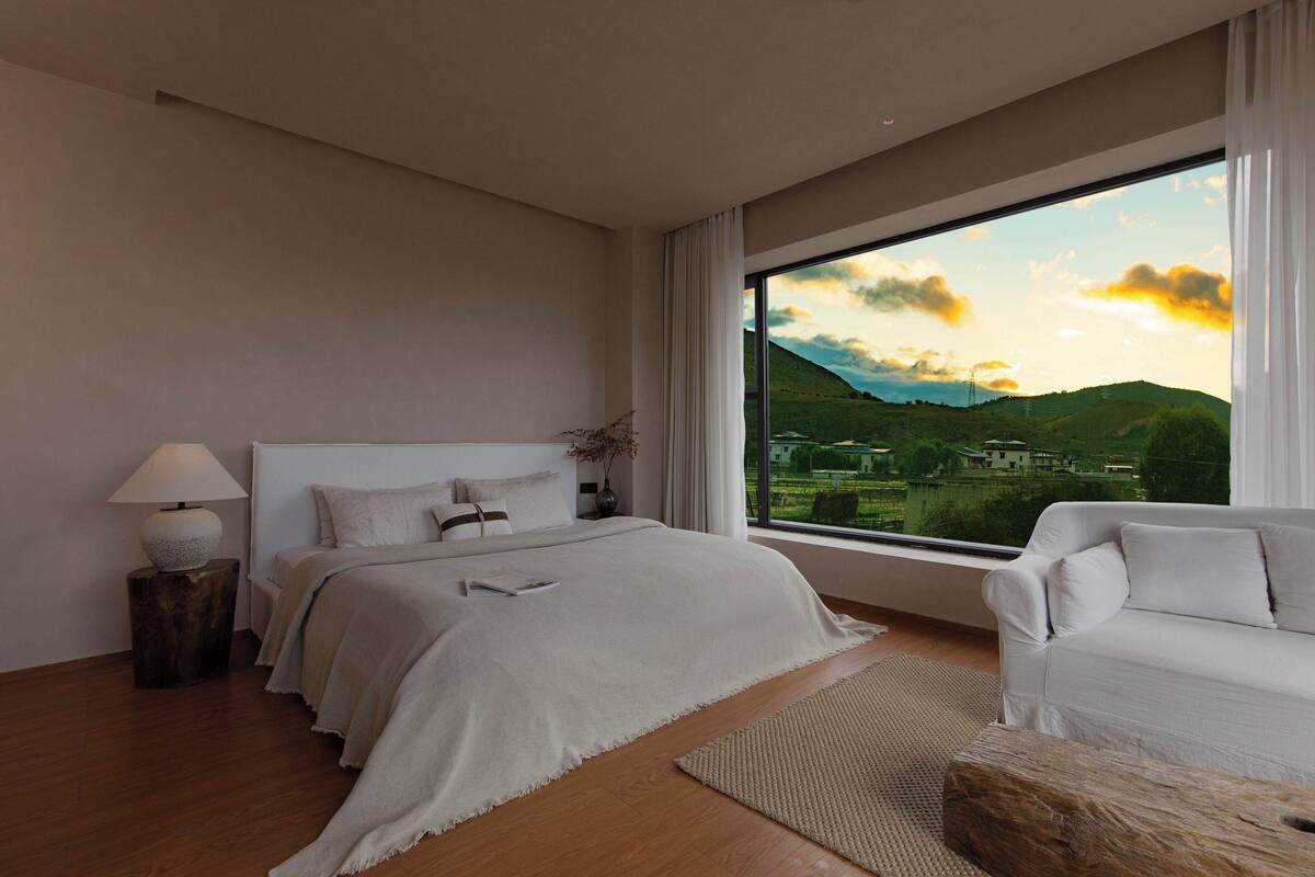 A modern bedroom with a large window, a bed, a bedside table with a lamp, and a sofa.