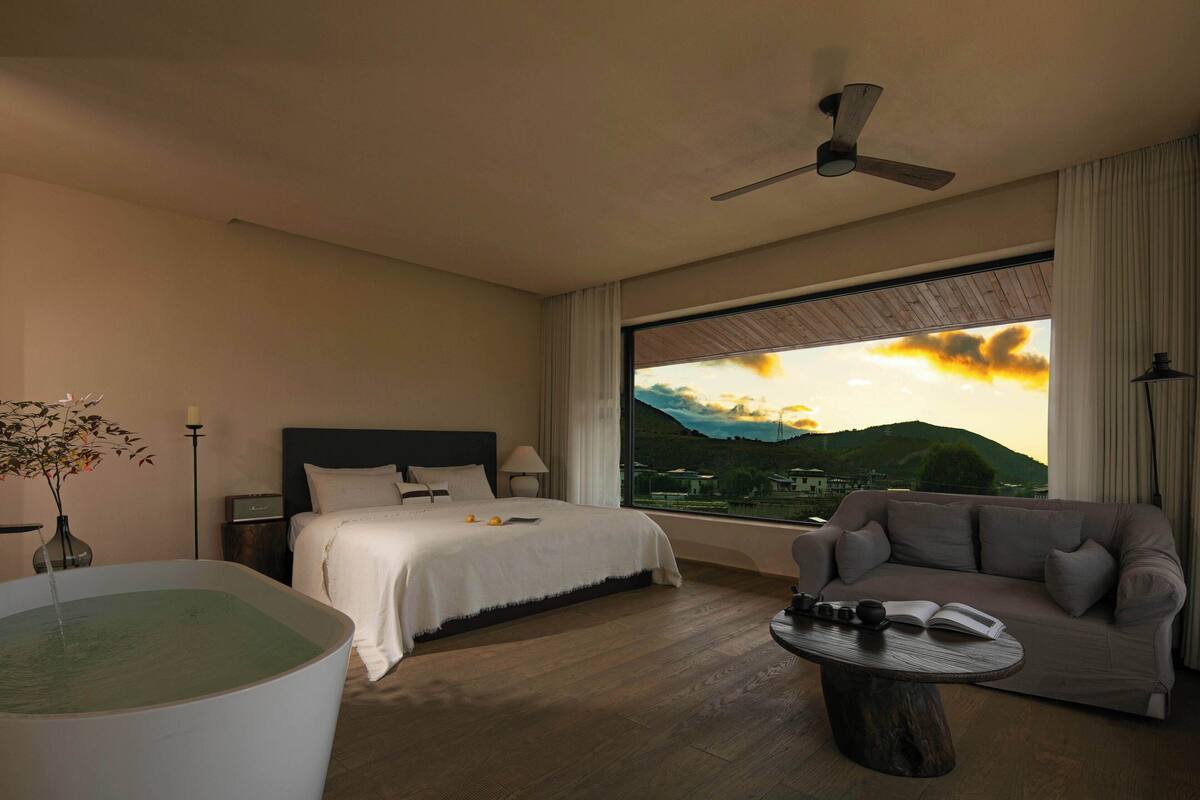 A hotel room with a bed, a sofa, a table, a large window with a view of mountains, and a ceiling fan.