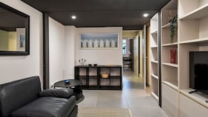 En moderne stue med en sort lædersofa, et lille rundt sofabord og et tæppe med et mønster. Der er et spiseområde med et sort konsolbord og et billede hængende over. Et fjernsyn er monteret på en hvid væg, og der er reoler med dekorative genstande.