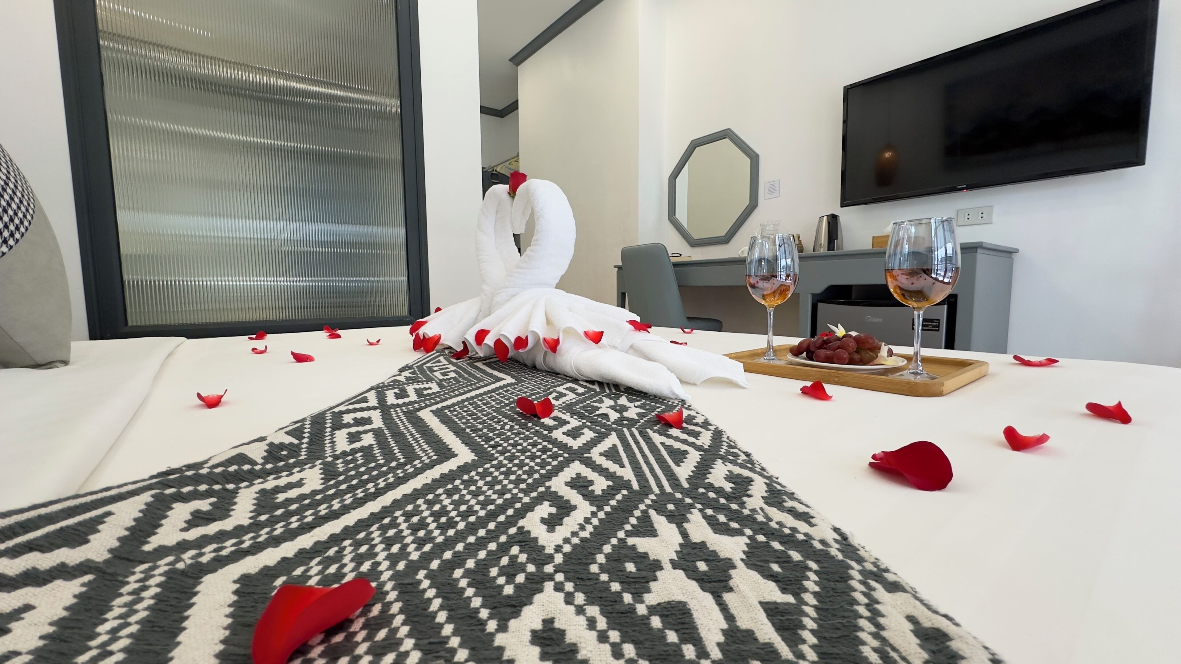 Una habitación de hotel con una cama bien hecha, un área de comedor con una bandeja de uvas y dos copas de vino, y un televisor montado en la pared.