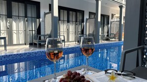 Escena junto a la piscina con dos copas de vino, un plato de uvas, un libro y una flor sobre una mesa.