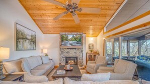 A living room with a stone fireplace, a ceiling fan, and a large window overlooking a deck. - Treetop Retreat in Connestee Falls (Brevard)