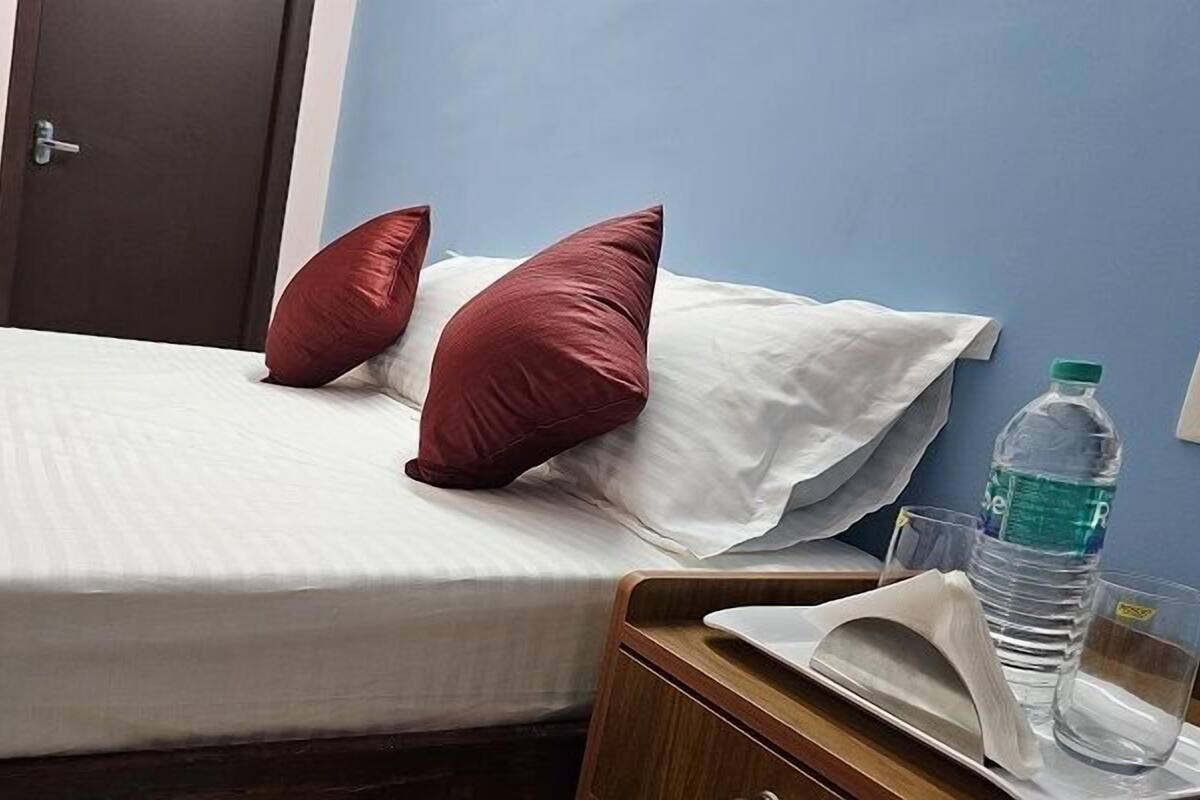 Habitación de hotel con cama, almohadas, mesita de noche con una botella de agua y vasos, y una puerta.