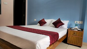 A neatly made bed with white and red bedding, a wooden nightstand with a tissue box and remote control, and a blue wall with a power outlet. - Pondy Pleasant Stay (Puducherry)
