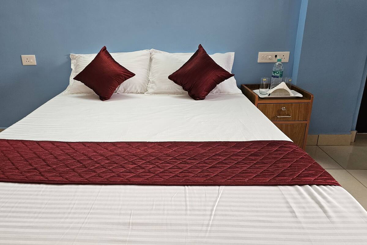 Una cama bien hecha con edredón blanco y dos almohadas rojas, una colcha roja y una mesita de noche de madera con una botella de agua y un vaso.