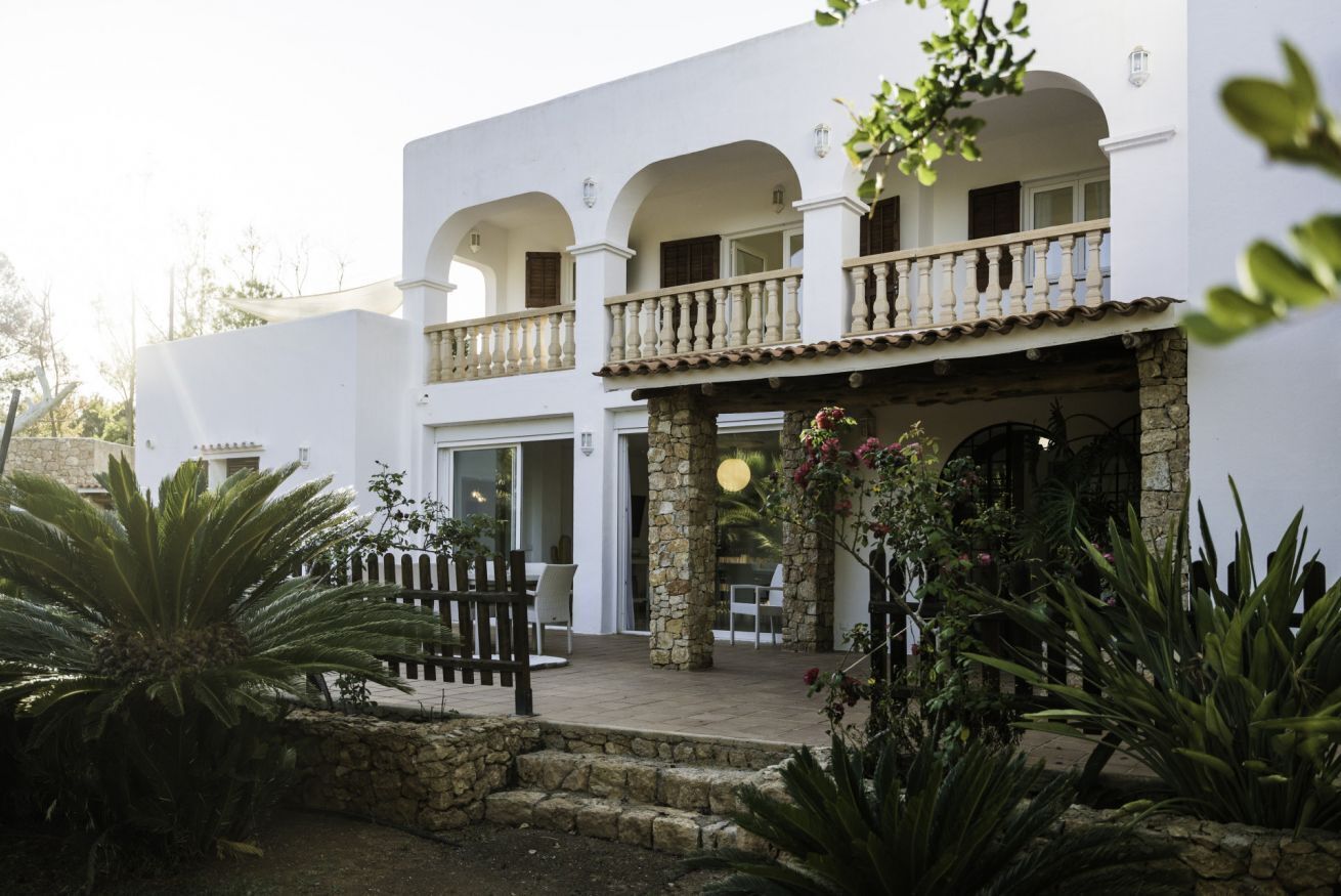 A white building with a balcony, stone pillars, and a garden with plants and a fence.