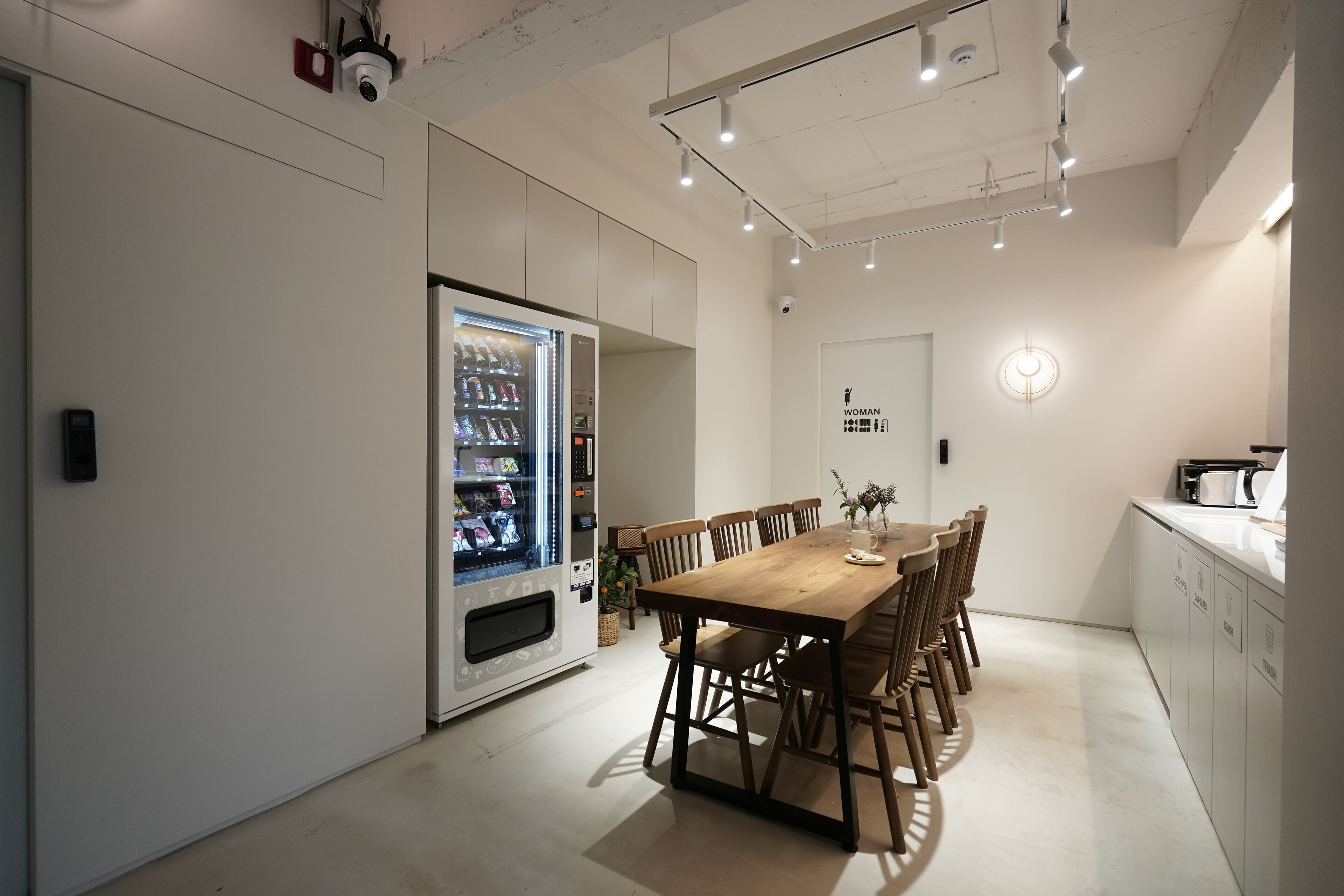 A modern dining area with a wooden table and chairs, a vending machine, and a kitchen area with a sink and cabinets.