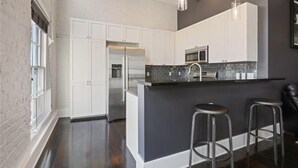 Una cocina moderna con armarios blancos, electrodomésticos de acero inoxidable y un salpicadero oscuro. Hay dos taburetes de bar y una ventana que proporciona luz natural.
