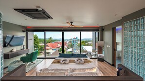A modern hotel room with a large bed, a TV, a green chair, and a view of the city. - Beautiful Sea View Full Smart House 3BR (Rawai)