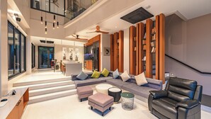 A spacious living area with a sectional sofa, ottomans, a leather armchair, and a coffee table. There is a staircase leading to an upper level, and a kitchen area is visible in the background. - Beautiful Sea View Full Smart House 3BR (Rawai)
