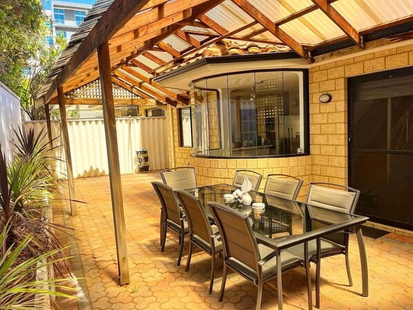 A covered outdoor patio with a glass table and chairs, a brick wall, and a window overlooking the area.