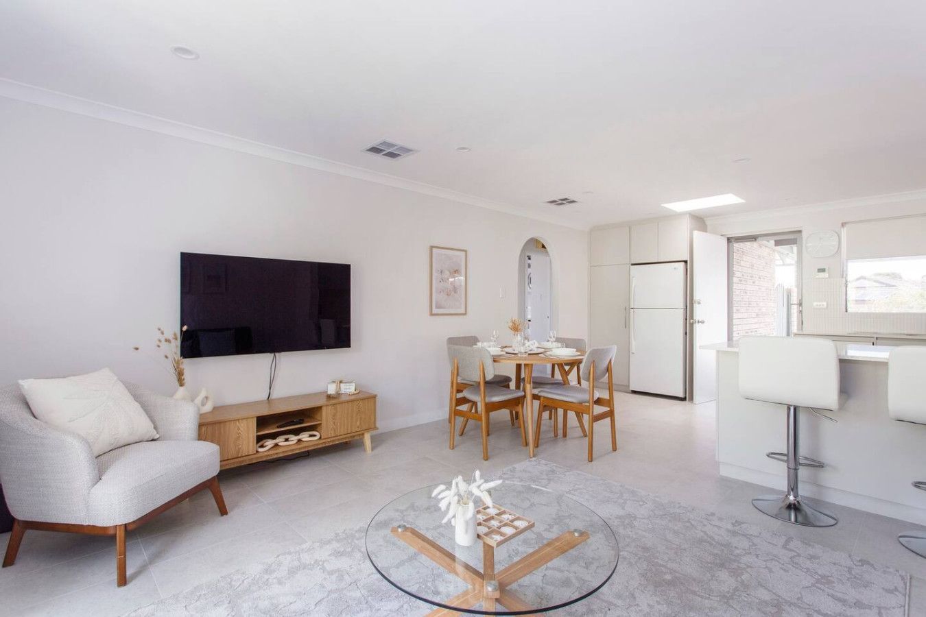 A modern living room with a flat-screen TV, a dining area with a table and chairs, a kitchen with a fridge, and a bar area with stools.