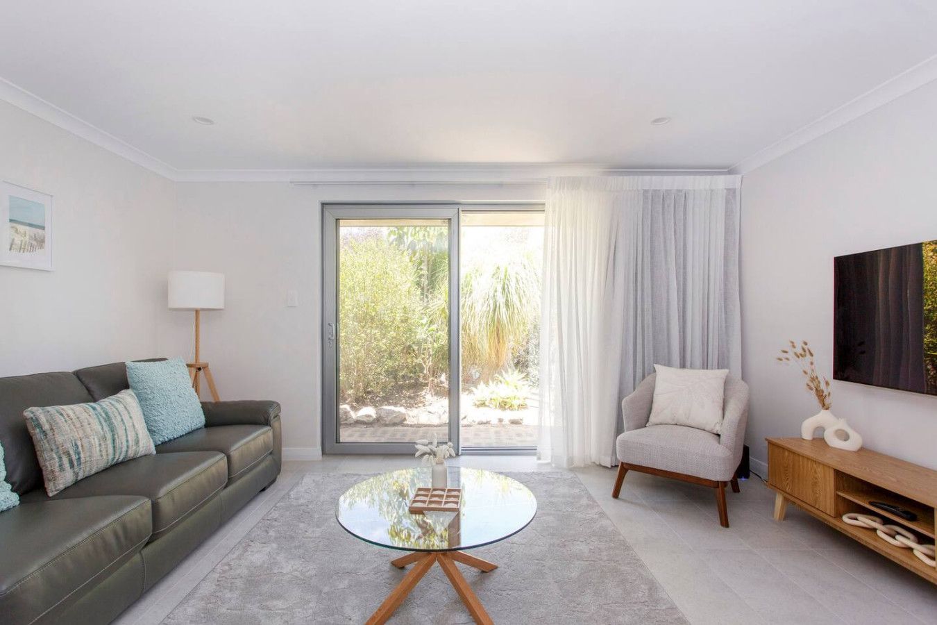 A modern living room with a sofa, armchair, glass coffee table, and a view through a sliding glass door to an outdoor area.