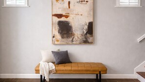 Una stanza con una panca in legno, un grande quadro astratto appeso alla parete e due finestre con tende bianche.