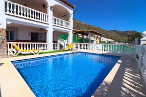 A swimming pool with a white fence, a balcony with yellow chairs, and a mountain in the background.