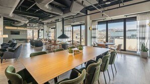 Une salle de conférence spacieuse, dotée d’une longue table en bois, de chaises vertes et de plantes en pot. De grandes fenêtres offrent une vue sur le paysage urbain et les montagnes.