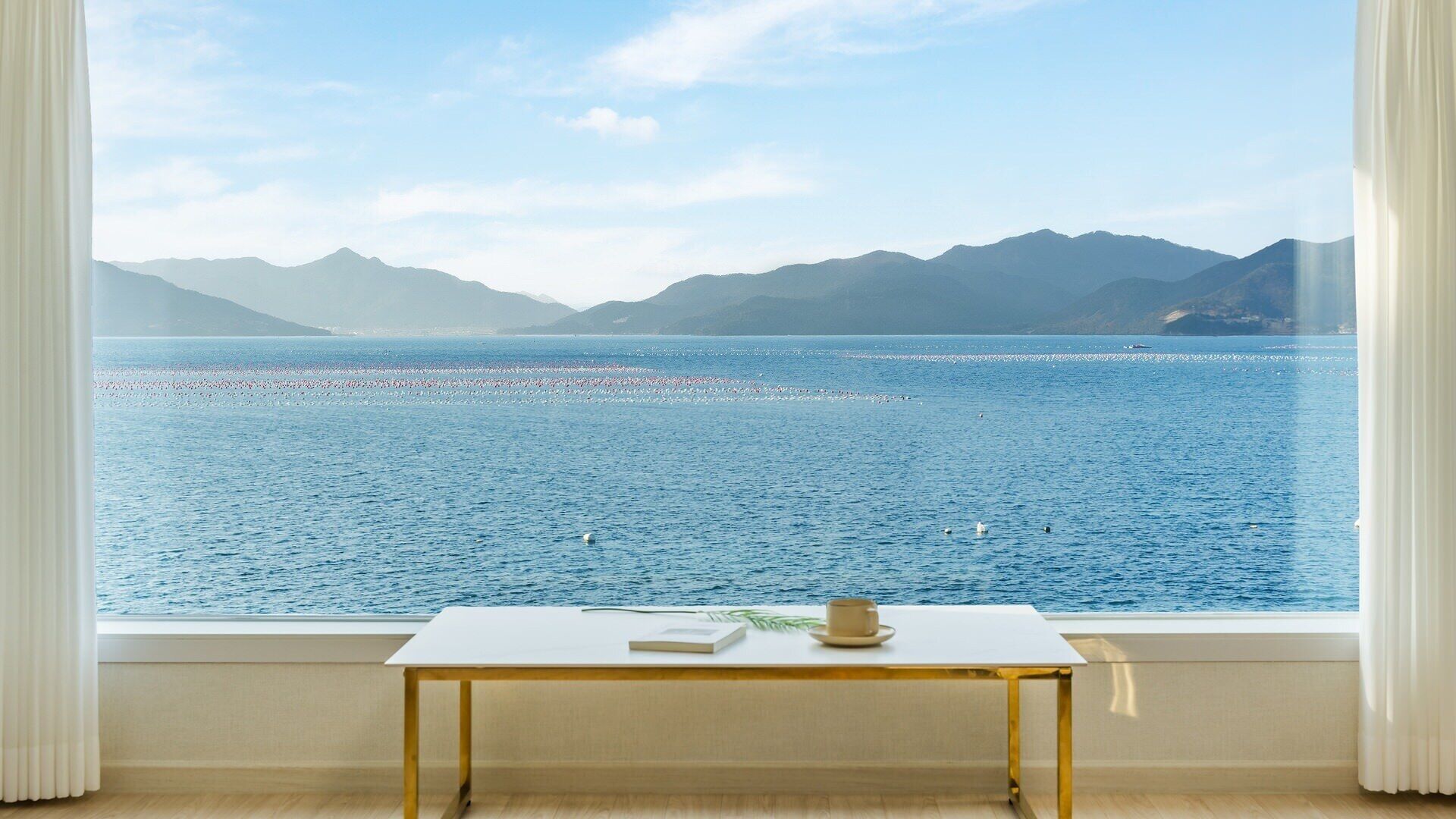 A room with a large window overlooking a calm sea and distant mountains.