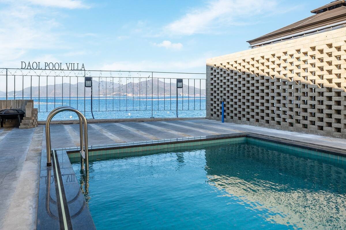 A swimming pool with a view of the sea, a sign reading "DAOL POOL VILLA", and a stone wall with a geometric pattern.