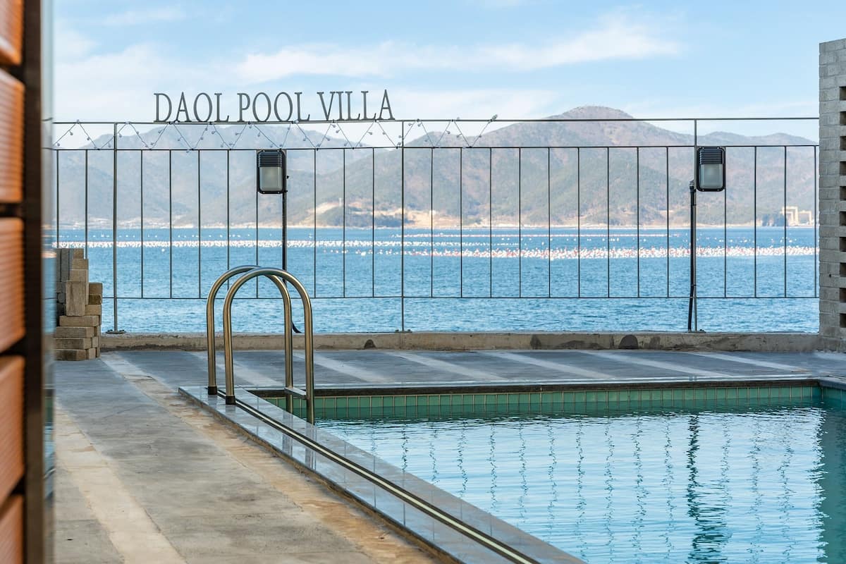 A pool area with a view of the sea and mountains, a metal fence, and a sign reading "DAOL POOL VILLA".