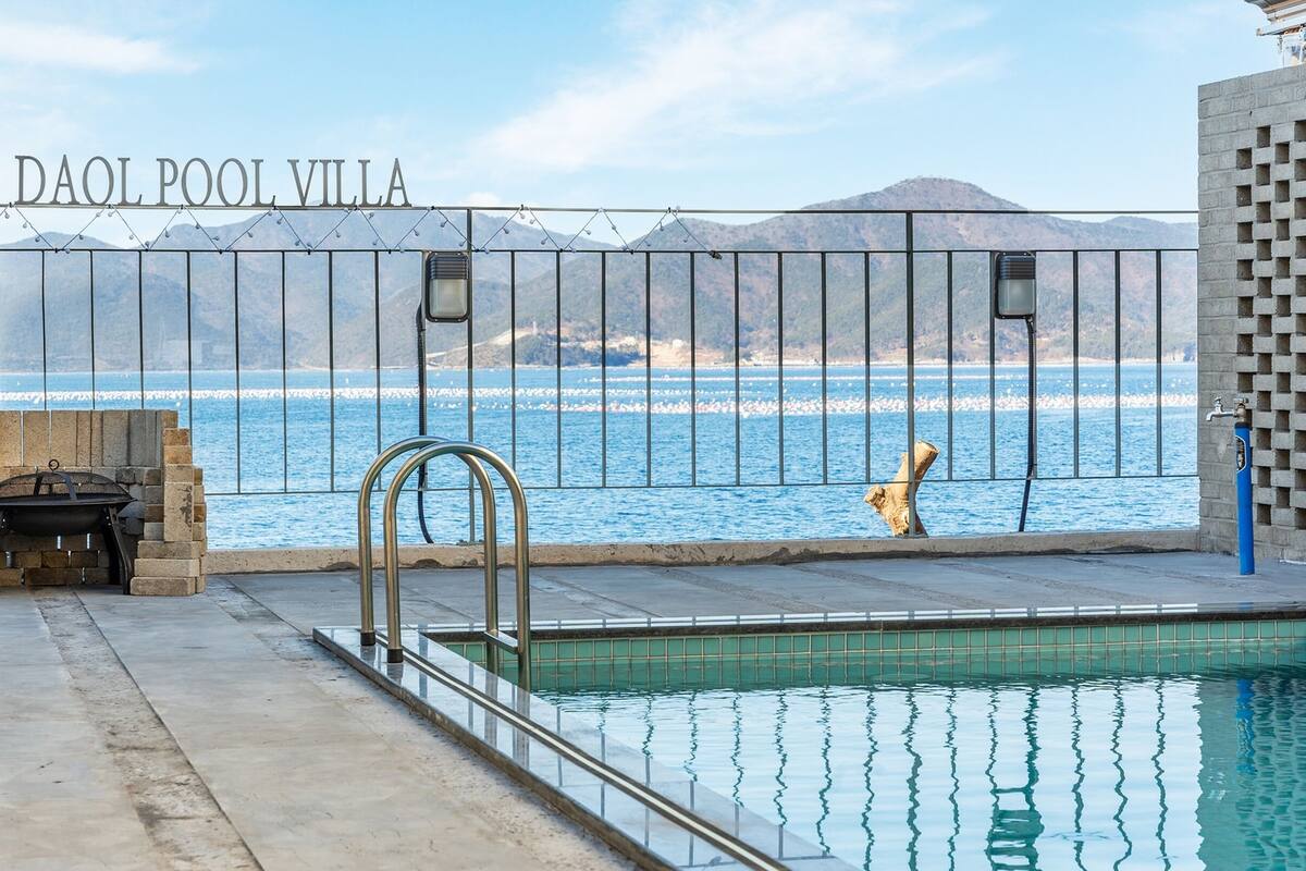 A pool area with a view of the sea and mountains, a sign reading "DAOL POOL VILLA", and a water fountain.