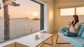 Una habitación con un ventanal grande con vista a un cuerpo de agua, una pequeña mesa con un pastel y un tenedor, y una persona sentada en un sofá azul.