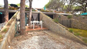 A stone-walled outdoor seating area with a table and chairs, surrounded by plants and trees.