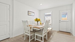 A dining area with a wooden table, white chairs, and a vase with sunflowers. There is a framed picture on the wall and a window with blinds.