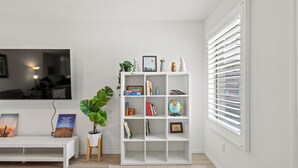 A modern living room with a white entertainment unit, a large flat-screen TV, a wooden bench, a potted plant, and a window with blinds.