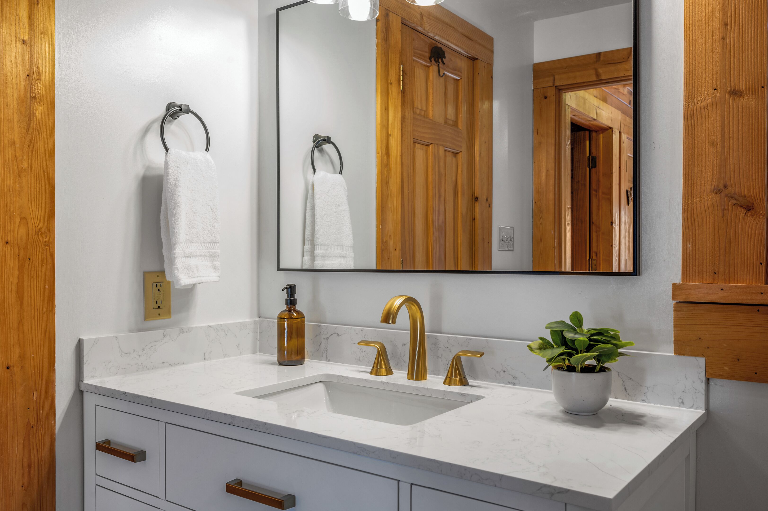 A bathroom with a white marble sink, gold faucets, a mirror, wooden cabinets, and a potted plant.