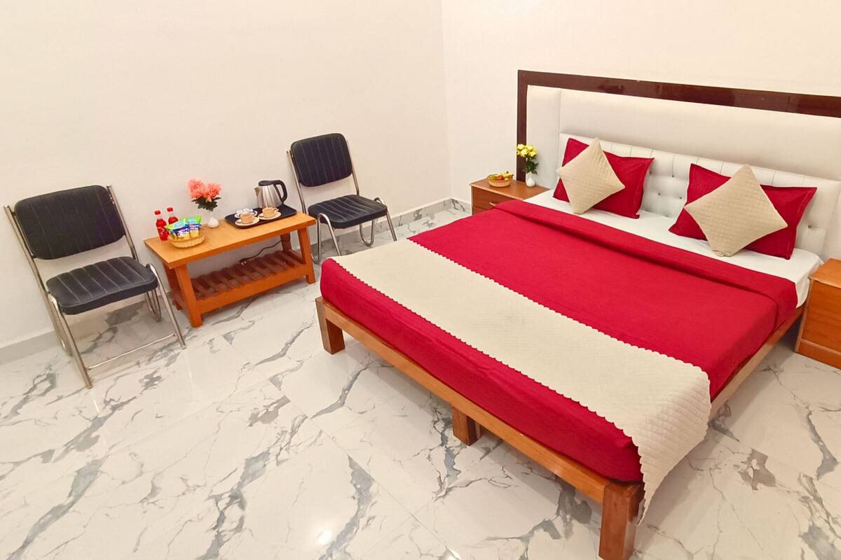 A hotel room with a bed, two chairs, a small table with a kettle, and a nightstand with a lamp and water bottles.