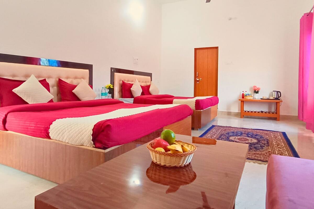 A hotel room with two beds, a wooden headboard, a small table with a fruit basket, a wooden side table with a kettle, and a door with a pink curtain.