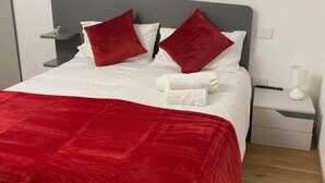 Sängyssä on harmaa pääty, valkoiset vuodevaatteet ja punaiset tyynyt.