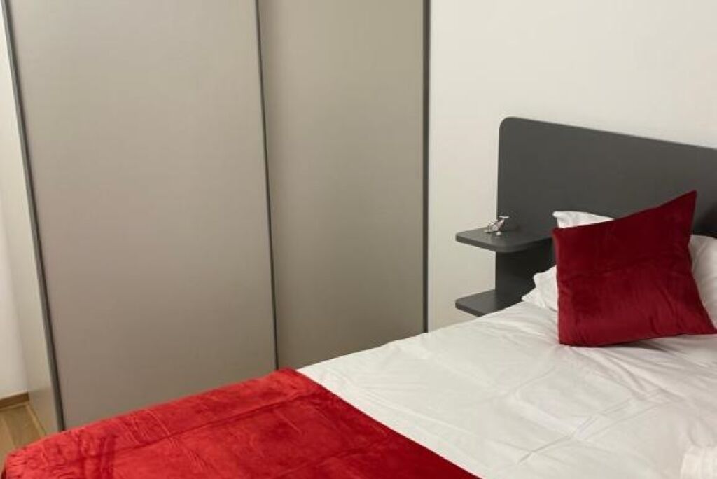 Una camera d'albergo con un letto, un comodino, una coperta a letto rossa e bianca e una testata del letto grigia.