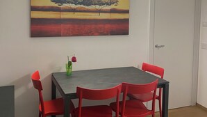 Ruokailutila, jossa on pöytä ja neljä punaista tuolia, pöydän yläpuolella oleva taulu ja oikealla puolella valkoinen ovi.