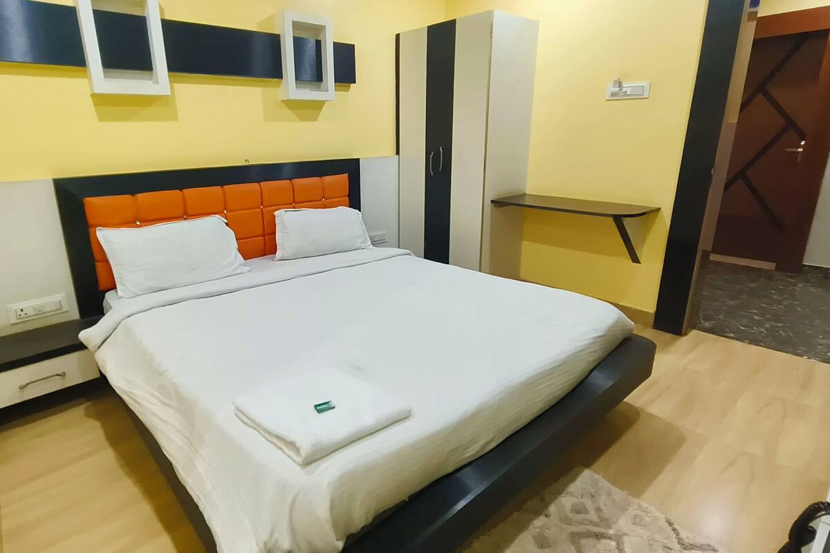 A neatly made bed with white linens and orange headboard pillows in a room with yellow walls, a wooden floor, and a small bedside table.