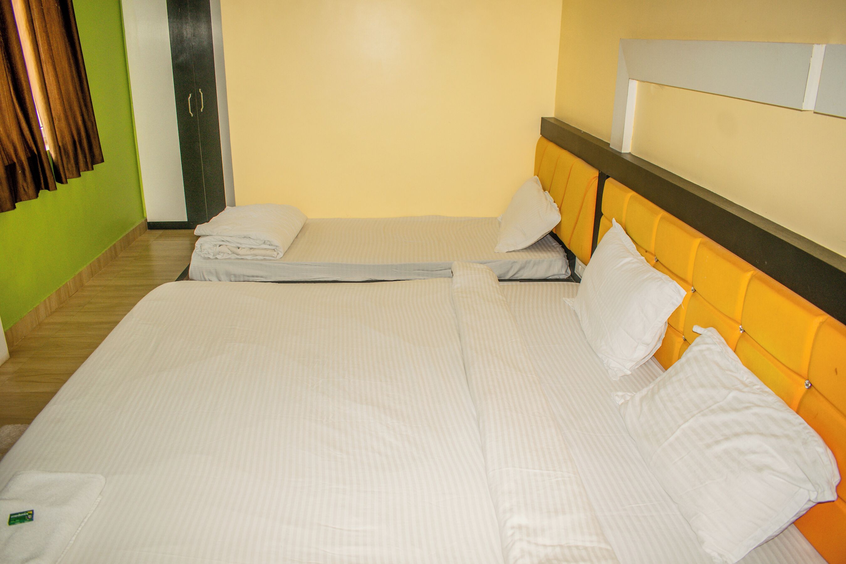 A double bed with white bedding and a yellow headboard, a green wall, and a wooden floor.