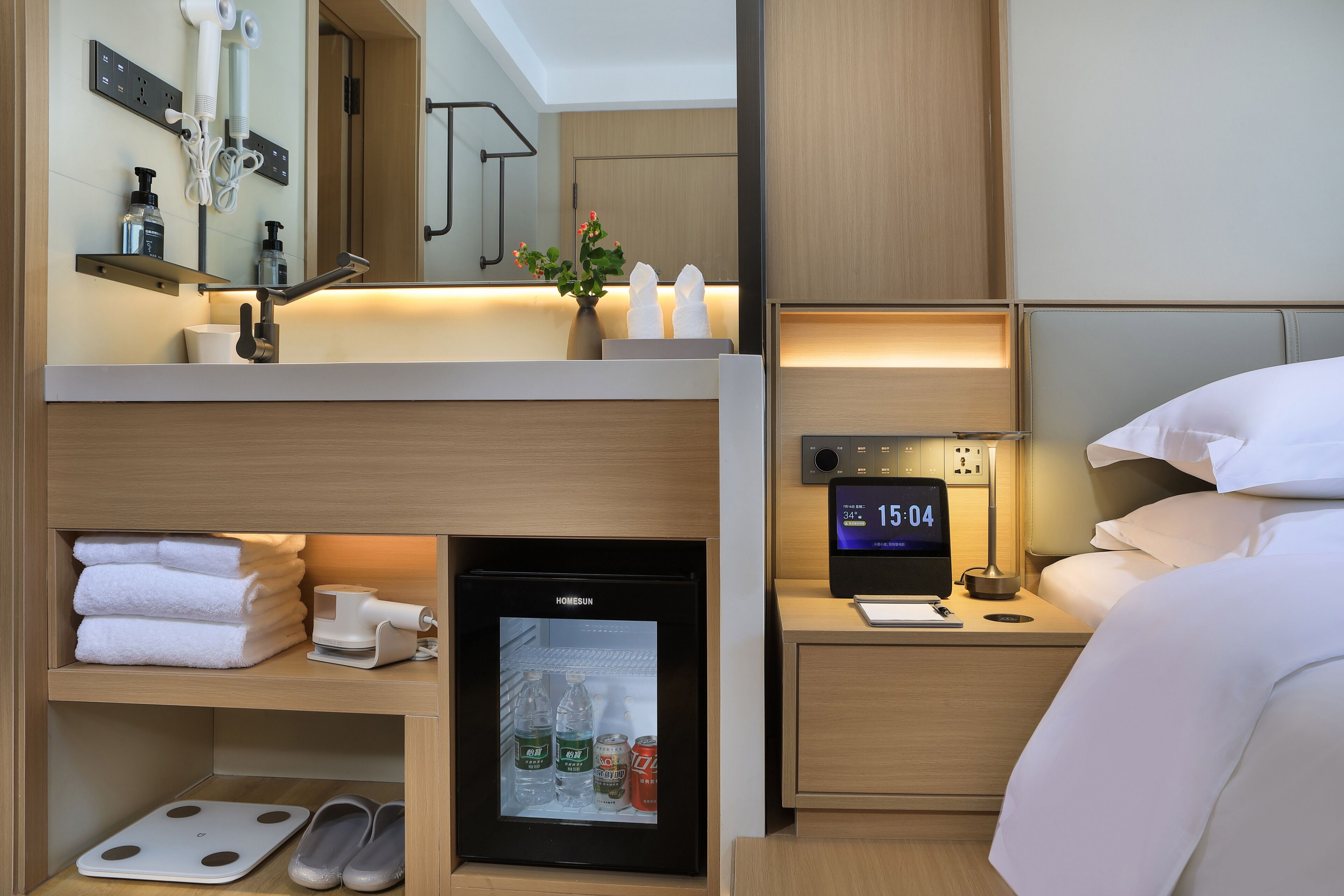 Habitación de hotel con cama, mesita de noche con un reloj, un mini refrigerador, un estante con toallas y una balanza.